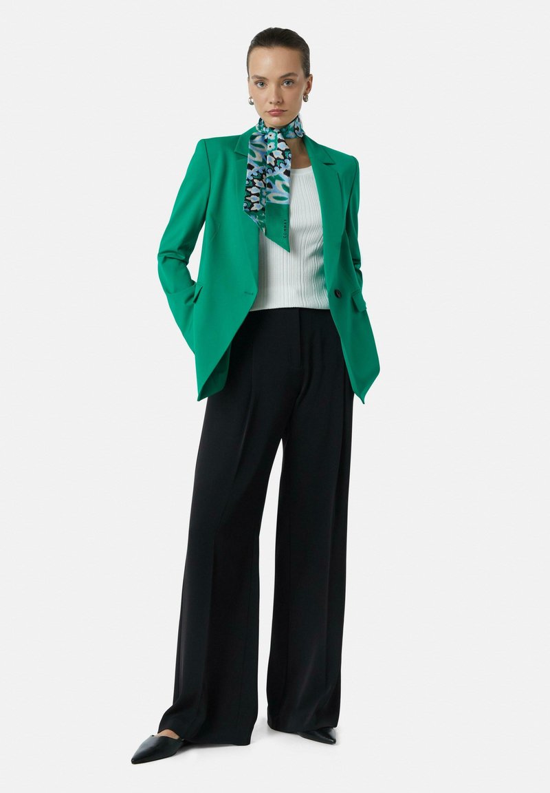 Groene blazer met een gestructureerd ontwerp, witte ribbeltop, zwarte wijde broek en een patroon sjaal met blauwe en groene accenten.