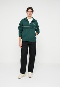 Zelená zipová bunda s bílými pruhy, slim fit, nošená přes bílou tričko; spojená s černými kalhotami a světlými teniskami.