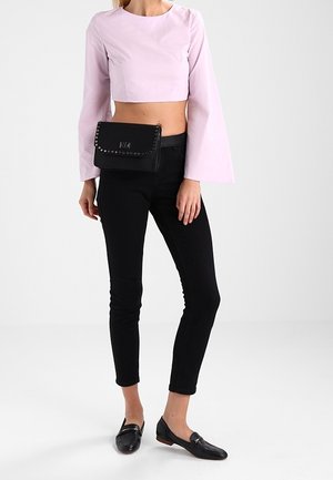 Mujer que lleva un top corto de manga larga color rosa claro, pantalones ajustados negros, mocasines negros y una riñonera negra con tachuelas.