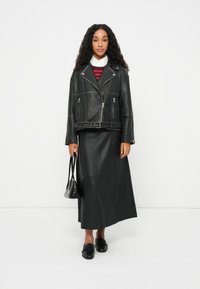 Μαύρο δερμάτινο μπουφάν μοτοσικλέτας με τσέπες που κλείνουν με φερμουάρ, ζώνη στη μέση και γιακά, συνδυασμένο με μακριά μαύρη δερμάτινη φούστα και μαύρα παπούτσια.