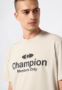 Hombre con camiseta beige con texto negro "Campeón Solo para Miembros" mira hacia su derecha contra un fondo claro y simple.