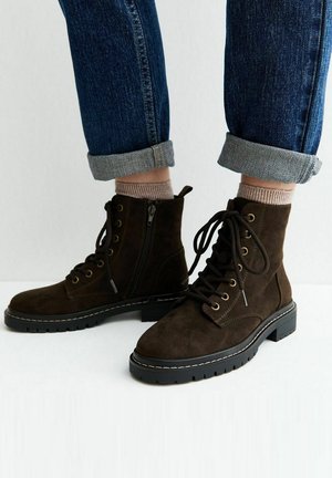 Botas de ante marrón oscuro con cordones y cremalleras laterales, llevadas con vaqueros azules remangados y calcetines beige sobre un fondo claro.