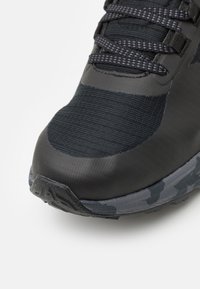 Baskets noires avec une tige texturée, comportant des zones lisses et striées, et une semelle à motif camouflage. Les lacets sont gris foncé.