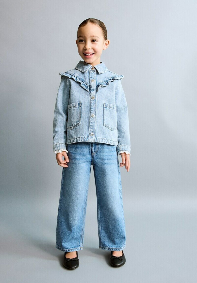 Fille souriante debout portant une veste en jean bleu clair à volants et un jean à jambes larges, chaussures noires, sur un fond gris uni.