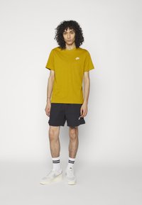 Rumena bombažna majica s belim logotipom Nike je kombinirana s črnimi kratkimi hlačami. Obutev so sivi čevlji Nike, poudarjeni z belimi in črnimi črtastimi nogavicami.