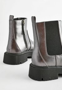 Bottines métallisées argent avec un panneau latéral élastique noir nervuré, semelle en caoutchouc noire et fermeture éclair sur le côté pour un accès facile.