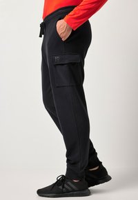 Osoba nosící černé cargo tepláky s boční kapsou a černé tenisky, ruka v kapse, a u pasu je vidět červený dlouhý rukáv.