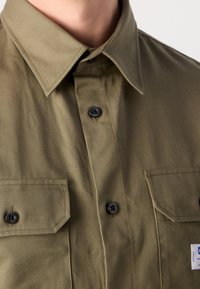 Gros plan sur une chemise vert olive avec des boutons noirs, un col pointu et deux poches poitrine à rabats boutonnés portée par une personne.