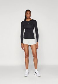 Melnas, garām piedurknēm sporta krekls ar apaļu kakla izgriezumu, kombinēts ar gaiši pelēkiem šortiem un baltām sporta kurpēm ar melnām svītrām.