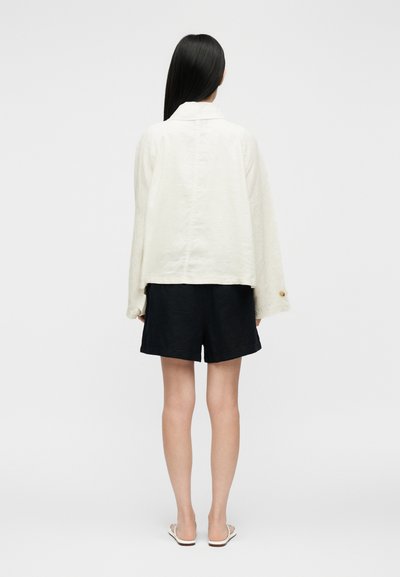 Žena s dlhými čiernymi vlasmi, oblečená v voľnej bielej blúzke s dlhým rukávom, čiernych krátkych nohaviciach a bielych sandáloch, stojaca chrbtom na jednofarebnom pozadí.