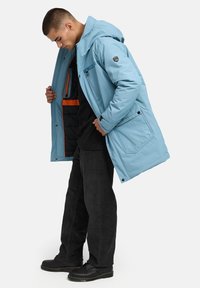 Gaiši zila, kapucēta jaka ar izšūtu oderi, priekšējām kabatām un oranžiem akcentiem. Saskaņota ar melnām biksēm un apaviem.