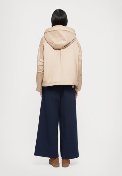 Veste matelassée beige avec capuche, présentant une silhouette courte. Associée à des pantalons larges navy et des chaussures à plateforme marron. Texture lisse.