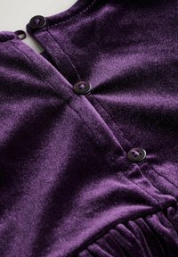 Purple velvet garment close-up featuring black buttons and a gathered texture along the hem, showcasing smooth fabric and detailed stitching.
