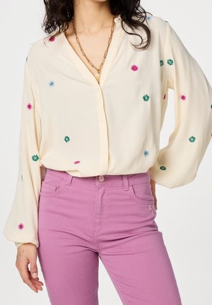 Femme portant un chemisier crème avec des fleurs brodées colorées et un pantalon rose taille haute, accessoirée d’un collier chaîne en or.