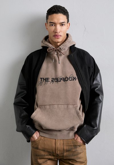 Schwarze Bomberjacke mit Lederärmeln, beige Kapuzenpullover mit aufgedrucktem Text und Kordelzug, kombiniert mit braunen strukturierten Hosen.
