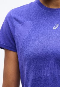 Κοντινό πλάνο ενός ατόμου που φοράει μωβ αθλητική μπλούζα Asics με υφασμένα σχέδια στο στήθος και το μανίκι.