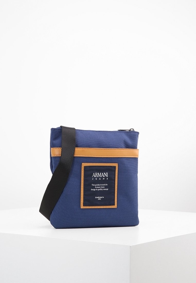 Granatowa torebka na ramię z płótna z brązowym skórzanym akcentem, zamknięciem na zamek i przednią etykietą. Posiada czarny, regulowany pasek.