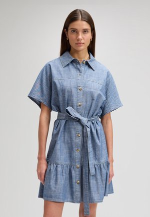 Mladá žena v chambray šatech na knoflíky s páskem, krátkými rukávy a volánkovým lemem, stojící před jednobarevným šedým pozadím.