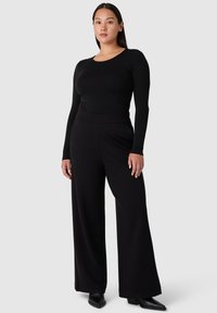 Musta pitkähihainen paita, joka on yhdistetty korkea vyötäröinen mustiin leveälahkeisiin housuihin, joissa on sileä pinta ja vartaloa myötäilevä siluetti. Mustat kengät viimeistelevät asun.