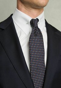 Krawatte mit Navy-Muster und geometrischem Design in Creme- und Rosttönen, kombiniert mit einem weißen Hemd und einem dunklen Sakko.