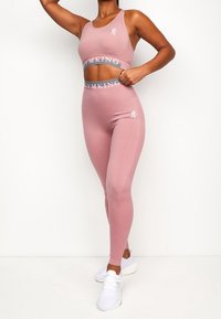 Růžová sportovní souprava zahrnující zkrácenou tílko a legíny s vysokým pasem, vyrobené z elastického materiálu s logem na pásu a bočními akcenty.