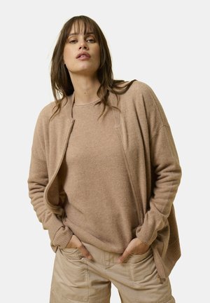 Beige Strickjacke mit strukturierter Oberfläche, offenem Design und gerippten Bündchen, über einem passenden, leichten Oberteil getragen, was einen harmonischen Look kreiert.