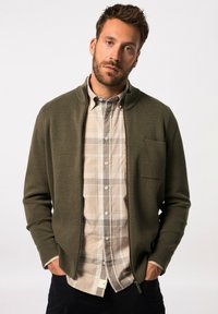 Suéter verde oliva con cremallera y cuello alto sobre una camisa de cuadros clara, que cuenta con un bolsillo en el pecho y una textura de tela.