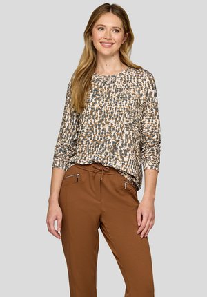 Langärmliges Oberteil in einem beigen und braunen Tierdruck, aus leichtem Material gefertigt. Kombiniert mit braunen Hosen mit Reißverschlusstaschen.