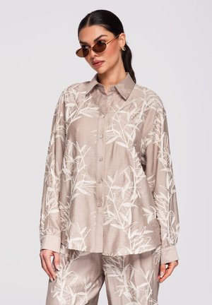 Vrouw draagt een oversized beige overhemd en broek met witte bladpatronen, bruine zonnebril, oorringen en haar naar achteren gebonden.