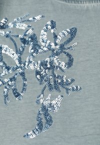 Zbliżenie srebrnych cekinów ułożonych w kwiatowy wzór na jasnoniebieskiej tkaninie.