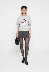 Szary sweatshirt z grafiką bałwanka i czerwonymi akcentami, w połączeniu z ciemną plisowaną spódnicą. Modelka nosi czarne rajstopy i czółenka.
