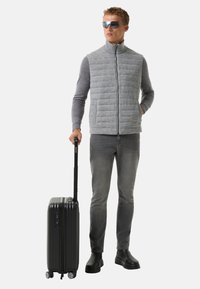 Graue gesteppte Weste über einem grauen Langarmshirt, dunkle Jeans und schwarze Schuhe, hält einen schwarzen Koffer mit ausziehbarem Griff.