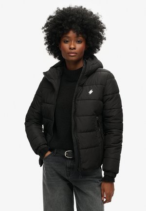 Schwarze Steppjacke mit Kapuze, ausgestattet mit einem Reißverschluss, elastischen Bündchen und einem kleinen weißen Logo. Wird über einem schwarzen Pullover und grauen Jeans getragen.