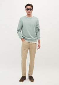Lys grøn crewneck sweatshirt, beige bukser, brune sko. Mand står afslappet med hænderne i lommen og iført sorte solbriller. Enkelt design, ingen mønstre.