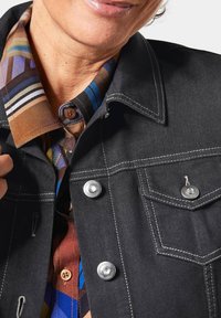 Schwarze Jeansjacke mit silbernen Druckknöpfen, weißen Nähten und Brusttasche, kombiniert mit einem mehrfarbigen gemusterten Hemd in Braun, Blau und gestreift.