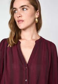 Chemise burgundy à manches longues avec un tissu texturé, des rayures verticales subtiles et un col rond. Présente une coupe ample et des boutons visibles.