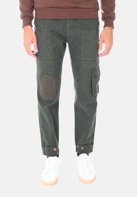 Pantaloni cargo verde oliva con molte tasche, toppe sulle ginocchia con cuciture rosse e orli regolabili. Abbinati a sneakers bianche.