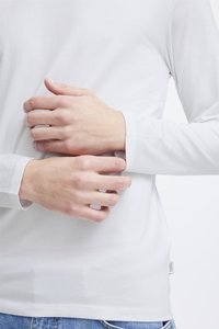 Fehér hosszú ujjú ing kerek nyakkal, sima textúrával, passzos mandzsettákkal és egy címkével az alsó oldalon, kék farmerrel párosítva.