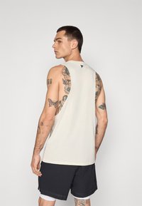 Light beige tank top with a rounded neckline and open armholes, featuring a small logo on the back. Paired with black shorts.