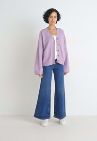 Cardigan oversized color lavanda con bottoni, abbinato a jeans in denim blu a zampa d'elefante e sneaker bianche. Tessuto morbido, vestibilità casual, design minimale.