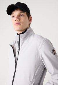 Ανοιχτό γκρι φούτερ με φερμουάρ και ψηλό γιακά, με μαύρο φερμουάρ. Διαθέτει λογότυπο στον γιακά και κυκλικό λογότυπο στο μανίκι. Φοράει μαύρο καπέλο.