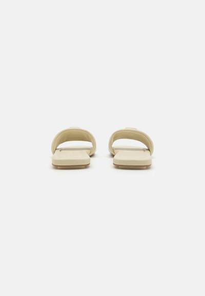 Beige slide sandals with a smooth material, open design, and flat sole. Includes a raised, rounded strap for support. No visible patterns or accents.