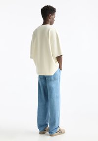 Sudadera de color crema, de gran tamaño y con mangas cortas, combinada con unos vaqueros de pierna ancha de color azul claro y zapatillas beige, vista desde atrás.