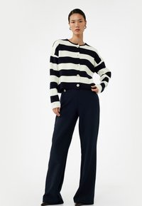 Cardigan a righe blu navy e bianche con scollatura rotonda e bottoni. Abbinato a pantaloni larghi scuri. Tessuto morbido e vestibilità su misura.