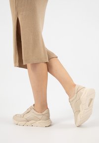 Sneaker atletici beige con tessuto texturizzato e dettagli lisci, caratterizzati da una suola spessa e pannelli laterali decorativi. Orlo a costine della gonna visibile.