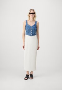 Denim crop top z gumbom, v kombinaciji z belim ribanim midi krilom in črnimi odprtimi natikači, postavljenimi na enobarvni ozadju.