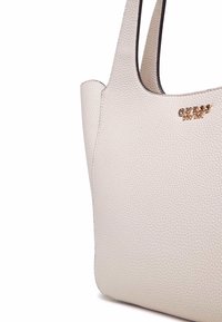 Borsa tote in finta pelle crema con finitura testurizzata. Presenta un'applicazione metallica con il logo "GUESS" sul davanti e due tracolle.