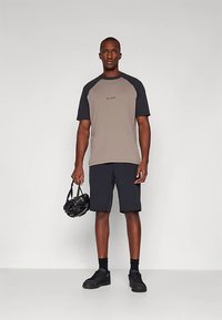 Άνδρας που στέκεται φορώντας πουκάμισο κοντομάνικο σε μπεζ και μαύρο, μαύρες βερμούδες, μαύρα αθλητικά παπούτσια, κρατώντας ένα μαύρο κράνος ποδηλάτου στο πλάι.