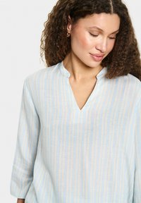 Blusa azzurro chiaro con strisce bianche verticali, caratterizzata da un colletto alla cinese e maniche a 3/4. Realizzata in un morbido tessuto testurizzato.