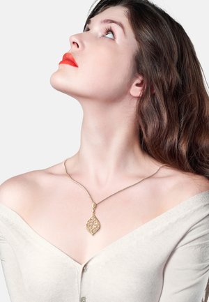 Goldener Anhänger in Form eines filigranen Blattdesigns, aufgehängt an einer dünnen Kette. Modell trägt einen schulterfreien, hellen Pullover, das Haar wellig.